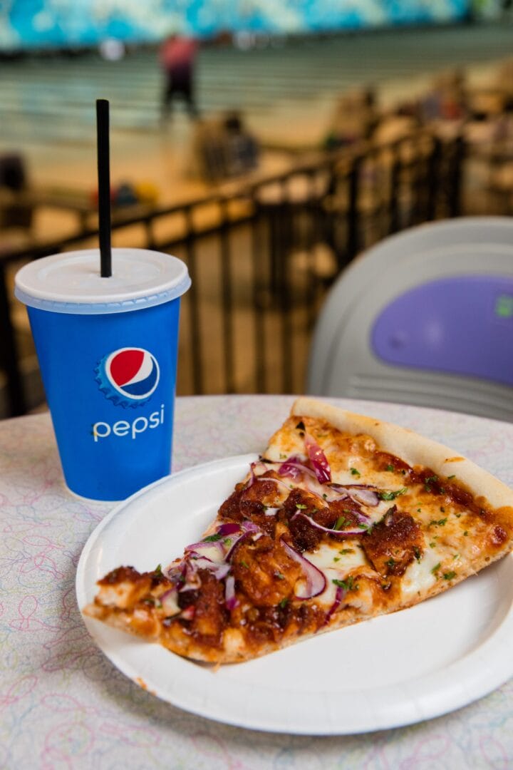 A slice of pizza with onions and chicken on a white plate next to a blue Pepsi cup with a straw, with a bowling alley in the background.