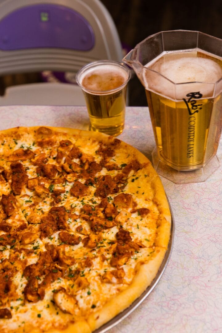 A whole chicken pizza on a table next to a glass and a plastic pitcher of beer, with a high chair visible in the background.