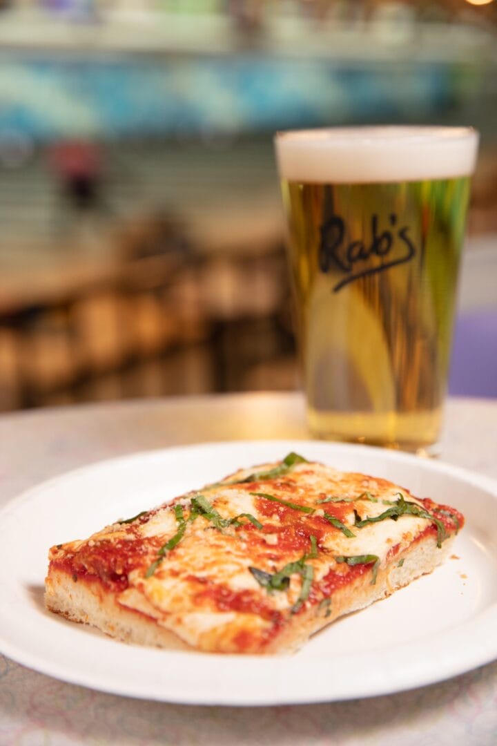 A slice of rectangular pizza with cheese and basil on a plate next to a pint of beer in a glass labeled "Rab's" on a table.