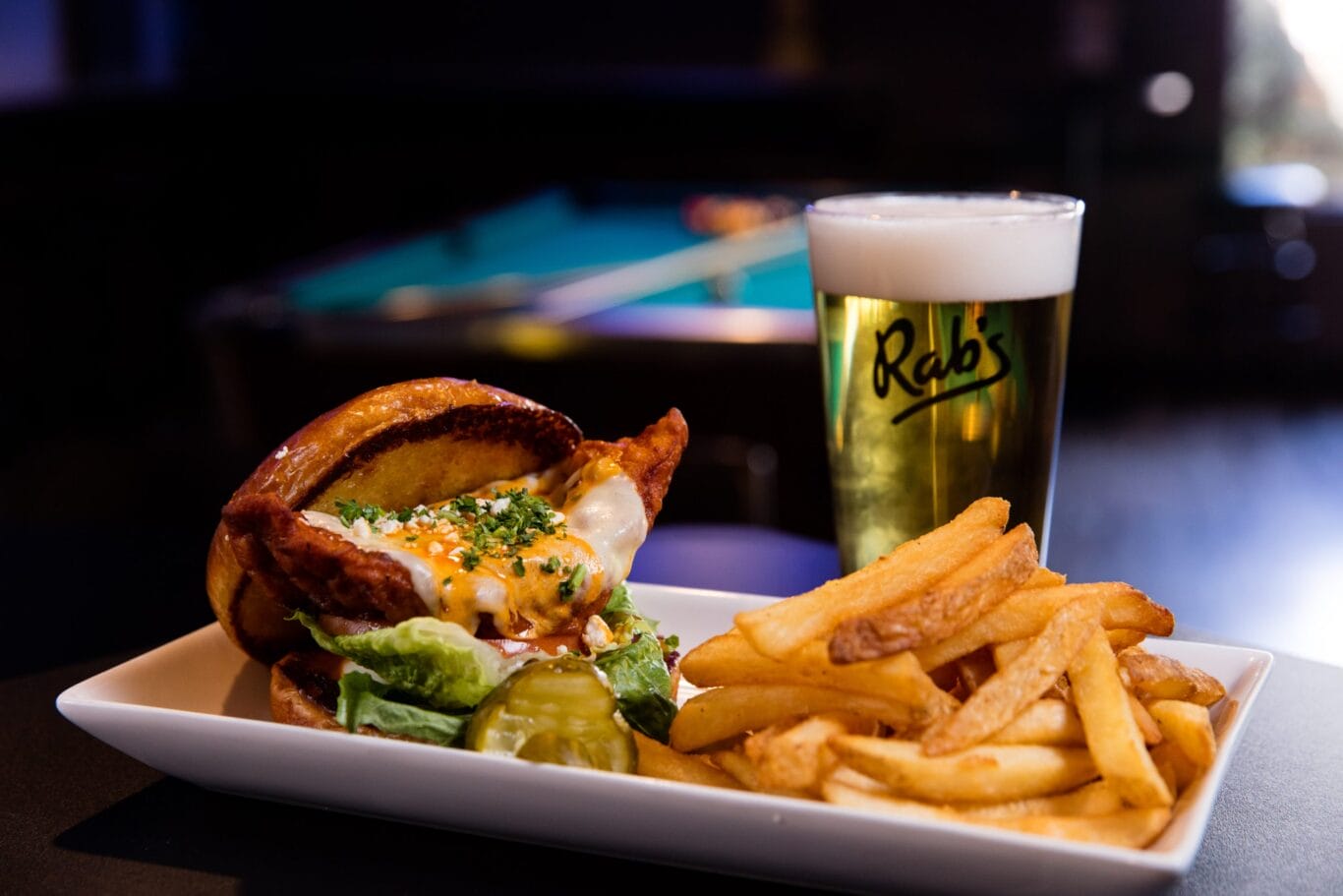 A plate with a cheeseburger, French fries, and pickles is next to a glass of beer on a table; a pool table is visible in the blurred background.