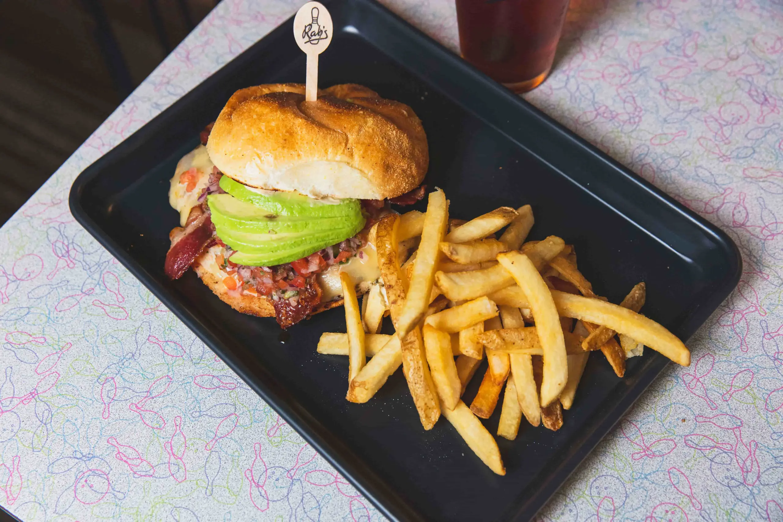 A hamburger with bacon, avocado slices, and cheese served with French fries on a black tray, next to a drink.