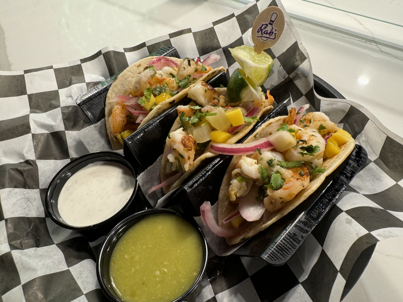 Three shrimp tacos with mango, onions, and cilantro, served on black holders with green salsa and white sauce, on black-and-white checkered paper.