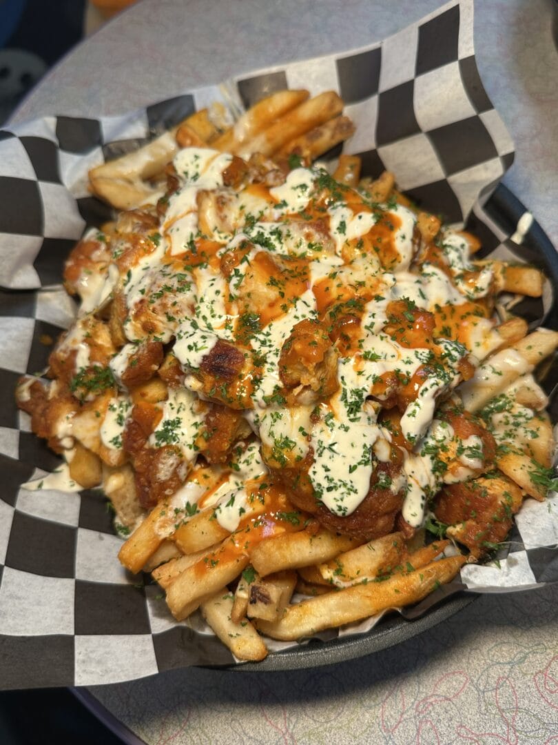 A basket of French fries topped with chopped chicken, creamy sauce, hot sauce, and chopped herbs, served on black and white checkered paper.