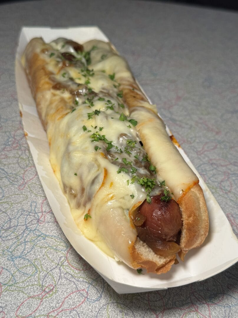 A hot dog in a bun is topped with melted cheese and sprinkled with chopped herbs, served in a white paper tray on a patterned surface.