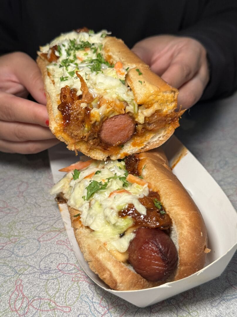 Two hands hold a hot dog topped with shredded cheese, slaw, and herbs above another similar hot dog in a paper tray on a patterned surface.