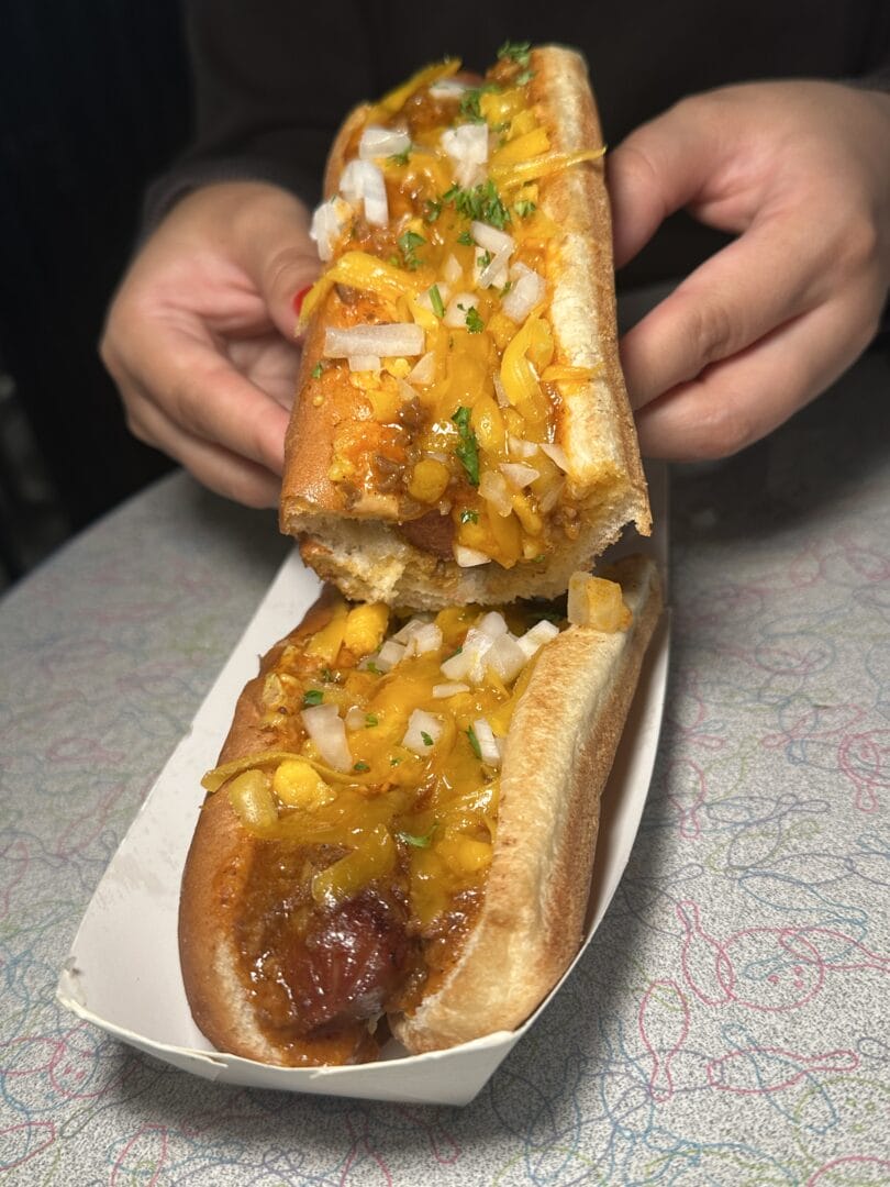 Two hands hold a hot dog topped with diced onions and yellow relish, while another chili dog rests in a paper tray on a patterned tabletop.