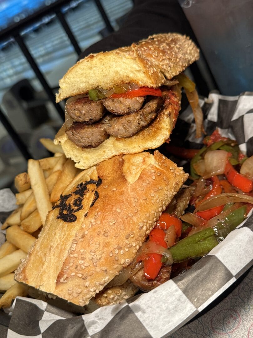 A sausage sandwich with grilled onions and peppers on a sesame roll, served with French fries on a checkered paper-lined tray.
