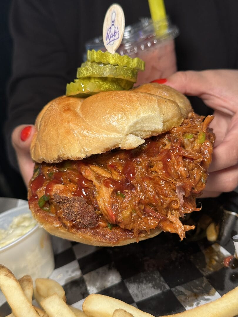 A person holds a pulled pork sandwich topped with barbecue sauce and pickles, with fries and coleslaw on the side.