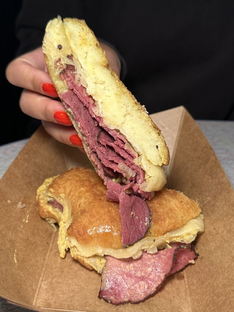 A person with red-painted nails holds half of a pastrami and cheese sandwich, with melted cheese and meat visible, over a brown paper tray.