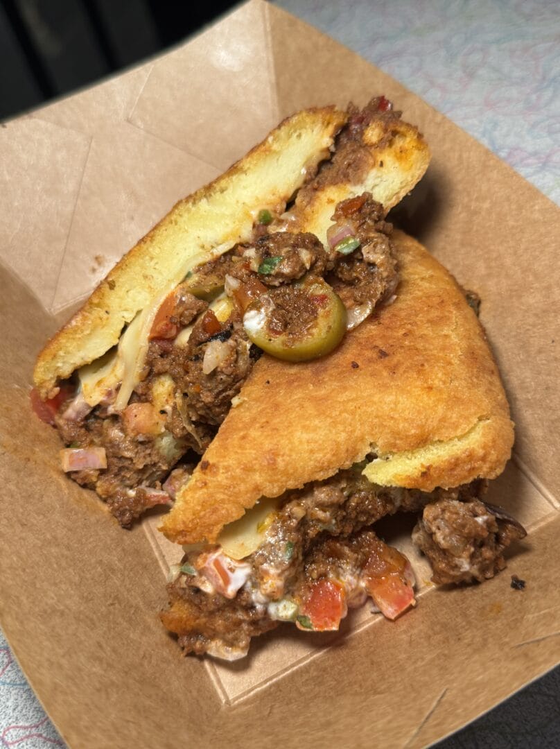 A close-up of a savory stuffed cornbread sandwich filled with seasoned ground beef, cheese, diced tomatoes, and topped with a sliced green olive, served in a brown paper tray.