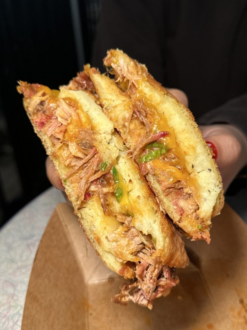 A hand holds a halved sandwich filled with shredded meat, melted cheese, and sliced peppers, served in a cardboard container.