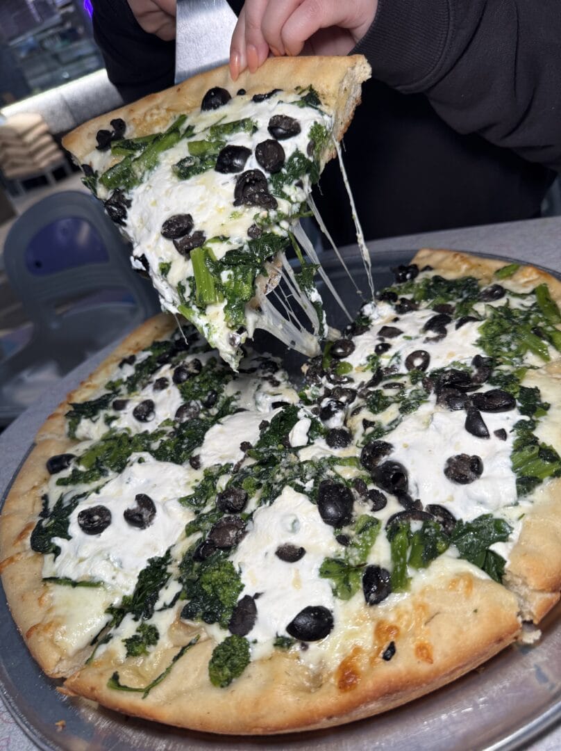 A hand lifts a slice of pizza topped with spinach, black olives, and melted cheese from a metal tray.