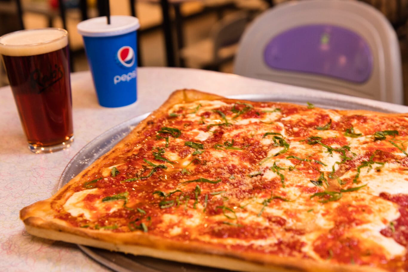 A large square pizza with tomato sauce, cheese, and green herbs sits on a table next to a glass of beer and a blue Pepsi cup.