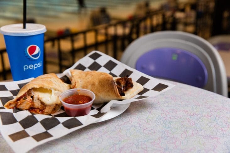 A tray with two stuffed calzones, a cup of salsa, and a Pepsi drink on a checkered paper, set on a table in what appears to be a casual dining area.