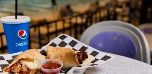 A tray with two calzones and a small cup of marinara sauce sits on a patterned table next to a Pepsi cup with a straw.