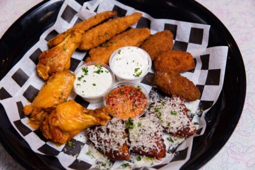 A black platter with chicken wings, breaded chicken tenders, mozzarella sticks, dipping sauces, and fried items on checkered paper.