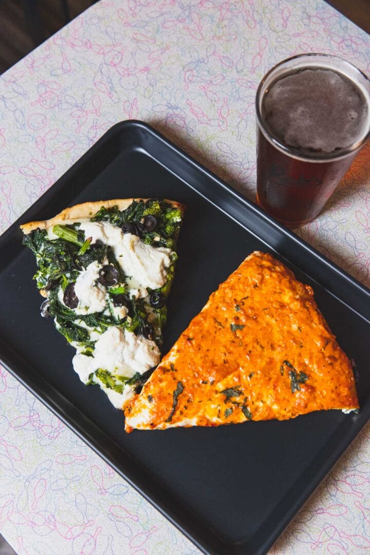 Two slices of pizza, one with spinach and ricotta, the other with red sauce, are on a black tray next to a glass of dark beer on a patterned table.