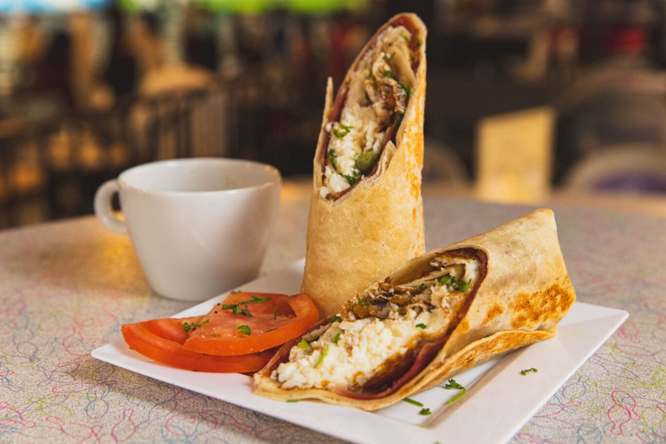 A wrap cut in half on a square plate with sliced tomatoes and herbs, next to a white cup on a table.