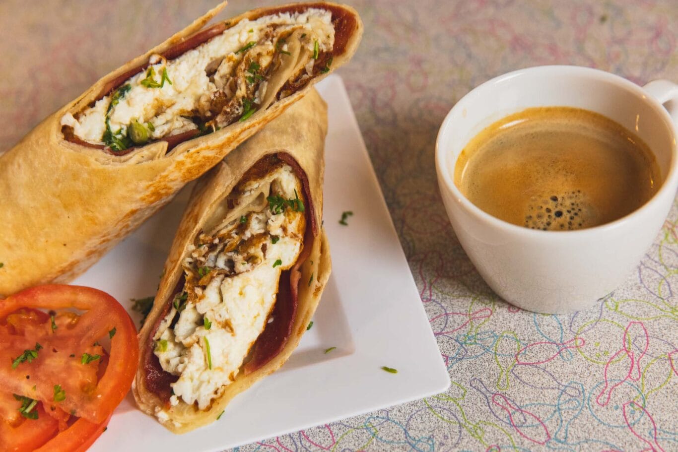Two halves of a stuffed wrap on a white plate with tomato slices, served next to a cup of coffee on a patterned tablecloth.