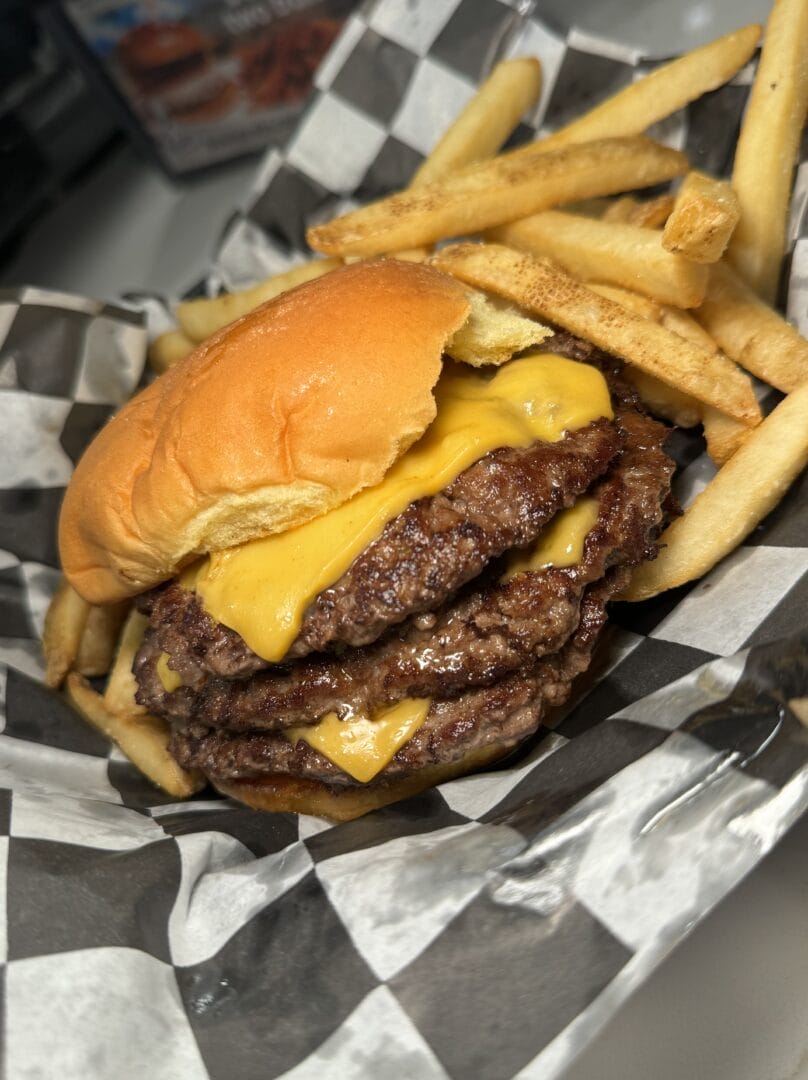 A triple cheeseburger with three beef patties and melted cheese, served with a side of French fries in a black and white checkered paper tray.