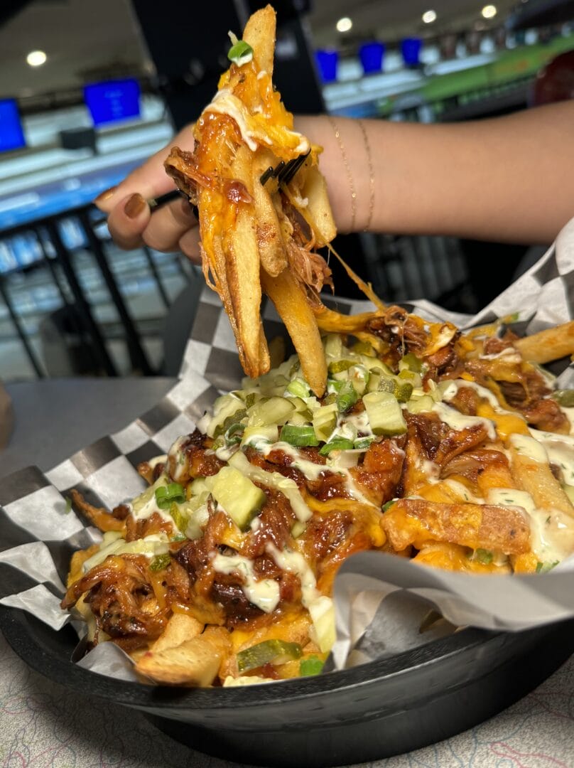 A hand lifts a portion of loaded fries topped with barbecue sauce, cheese, diced pickles, and chopped green onions from a black basket lined with checkered paper.