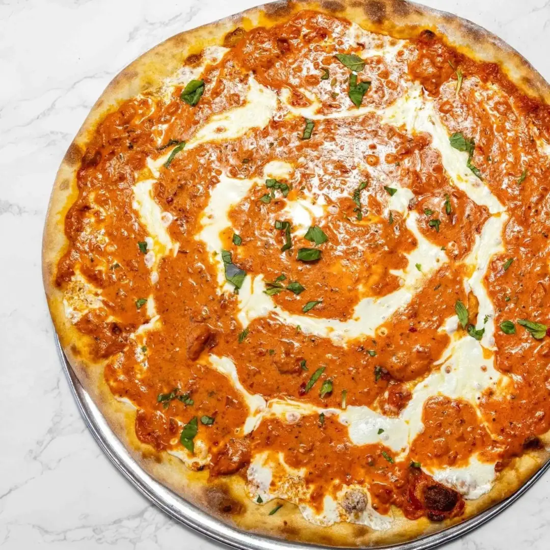 A whole pizza topped with a creamy orange sauce and scattered fresh herbs, served on a metal tray atop a marble surface.